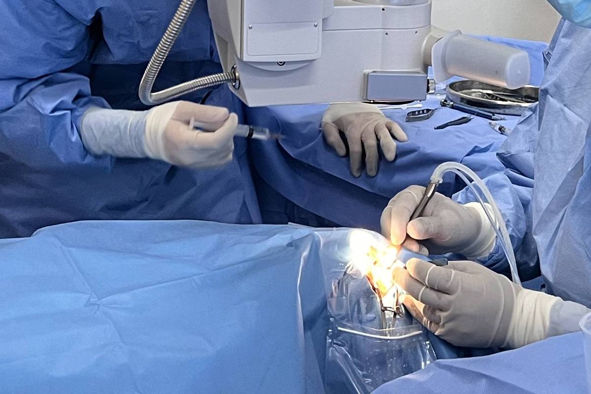 Três profissionais de saúde com roupas cirúrgicas e máscaras realizam cirurgia ocular. Dois estão operando com equipamento especializado, enquanto um observa ao lado.