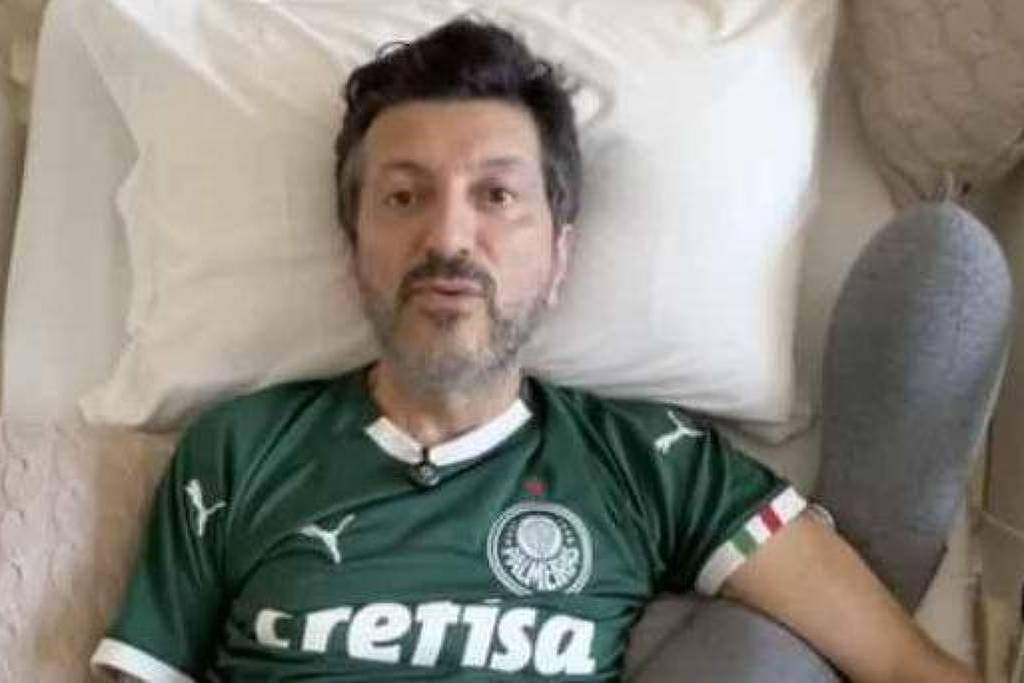 Homem de meia-idade deitado em cama branca, usando camisa verde do Palmeiras com logo da Crefisa. Travesseiro branco e almofada bege ao lado.