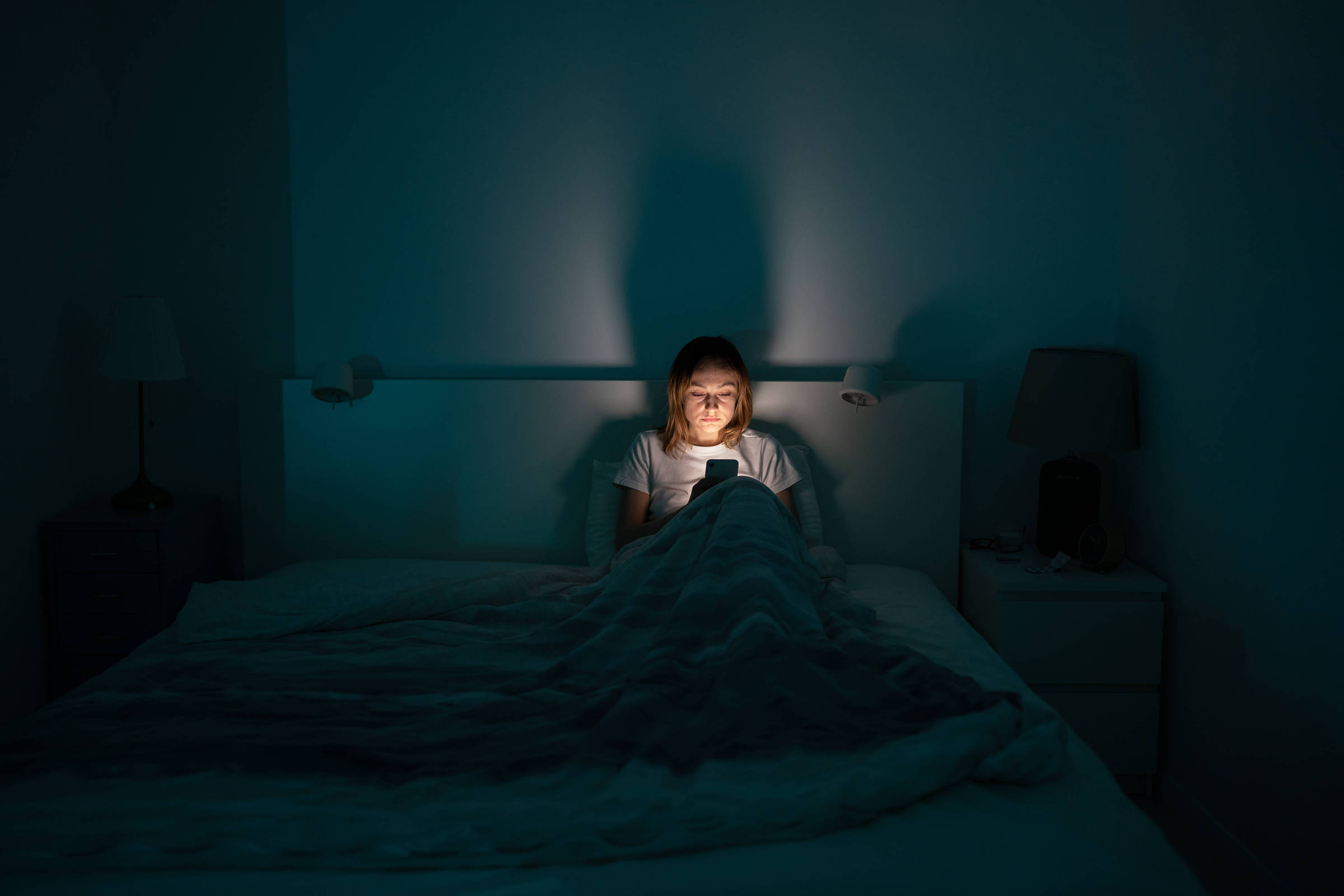 A imagem mostra uma pessoa sentada na cama, envolta em um cobertor, olhando para um celular que emite luz. O ambiente é escuro, com uma iluminação suave que destaca o rosto da pessoa. Ao fundo, há uma mesa de cabeceira com uma lâmpada acesa e uma outra mesa ao lado, também com um abajur. A sombra da pessoa é projetada na parede.