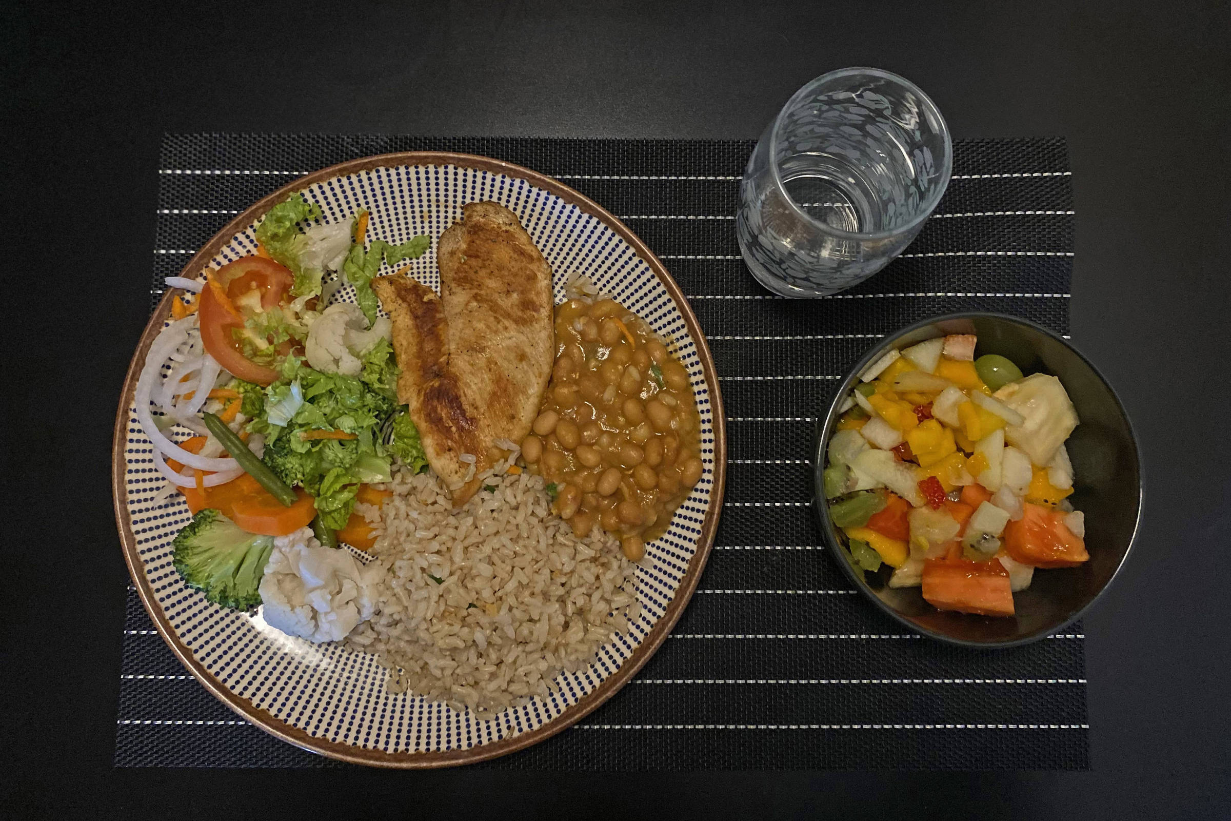 Prato contém filé de frango grelhado, feijão, arroz integral e salada com alface, tomate, cenoura, cebola e brócolis. Ao lado, tigela com salada de frutas variadas e copo com água sobre toalha preta com listras brancas.