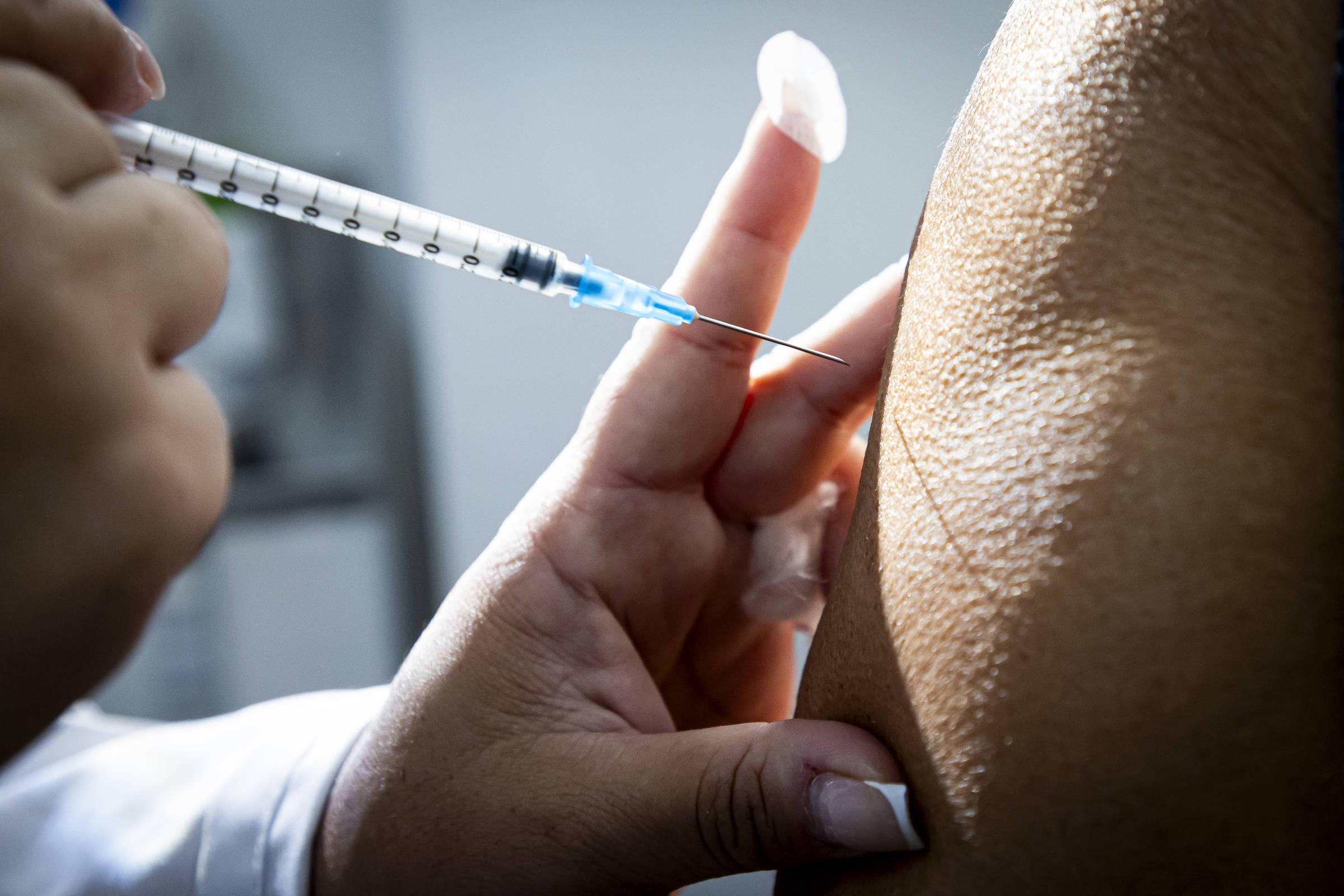 Mão de profissional de saúde segurando seringa próxima ao braço de pessoa para aplicação de vacina. Algodão branco está posicionado no dedo indicador do profissional.