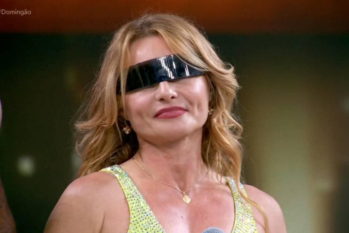 Mulher loira com cabelos soltos usa óculos pretos largos e brilhantes que cobrem os olhos. Ela veste vestido verde claro com brilho e segura microfone em estúdio com fundo desfocado.