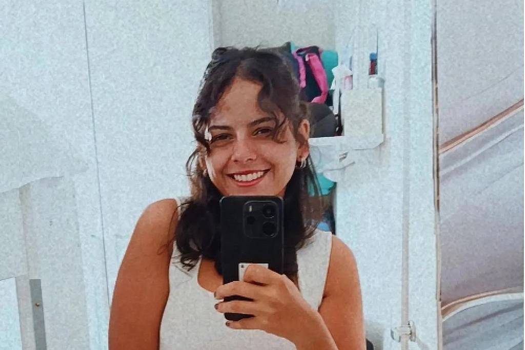 Mulher jovem com cabelo preso parcialmente, usando regata branca e calça listrada, sorri enquanto segura celular preto para tirar selfie em ambiente interno com paredes claras.