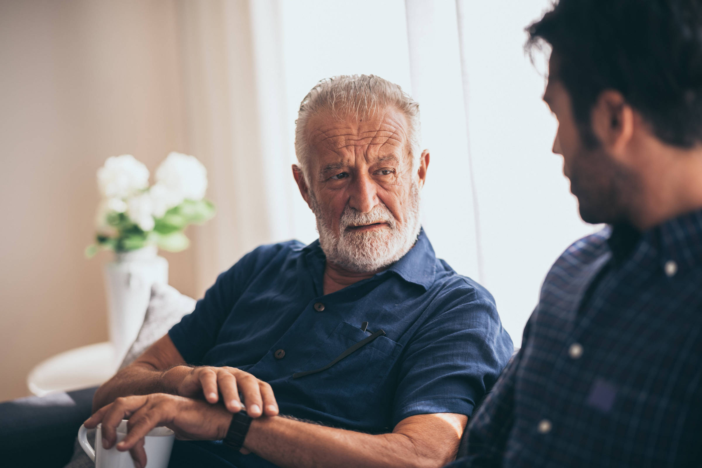 Homem idoso de cabelos grisalhos e barba branca veste camisa azul escura e conversa com homem mais jovem de camisa xadrez azul em ambiente interno iluminado por luz natural. Ao fundo, vaso branco com flores brancas desfocado.