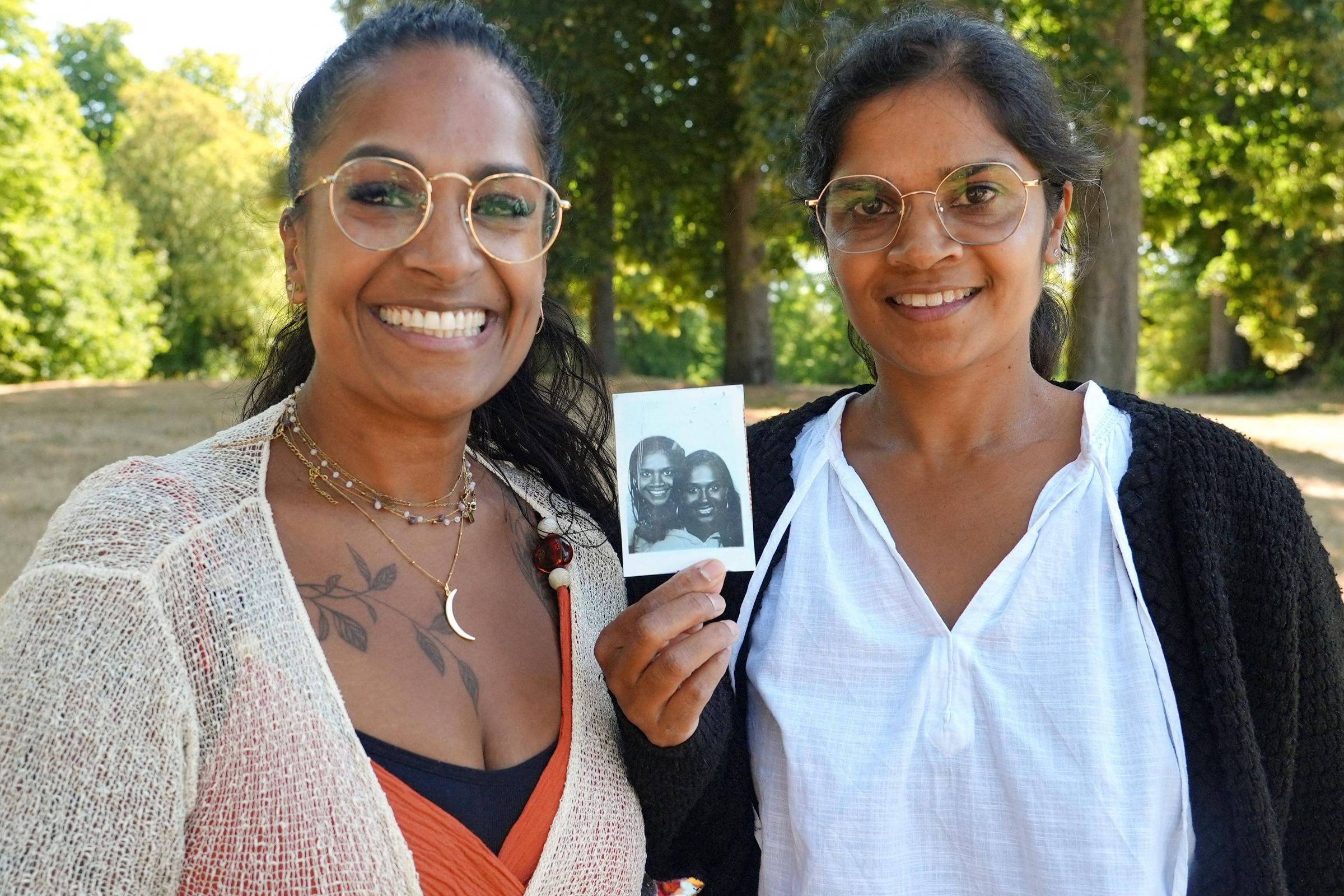 Duas mulheres com óculos estão em um parque com árvores e gramado seco ao fundo. A mulher à esquerda usa blusa clara transparente sobre roupa laranja e colares, e a da direita veste blusa branca com cardigã preto, segurando uma foto em preto e branco de ambas sorrindo.