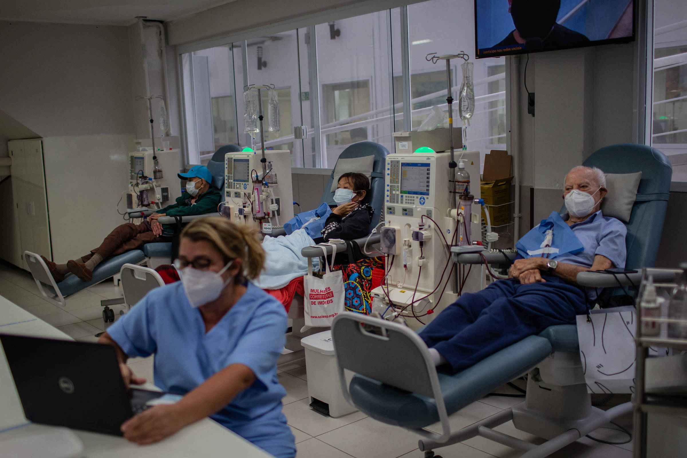 Três pacientes sentados em cadeiras reclináveis recebem hemodiálise em clínica. Todos usam máscaras faciais. Profissional de saúde em primeiro plano trabalha em computador. Equipamentos médicos e bolsas de soro estão visíveis.