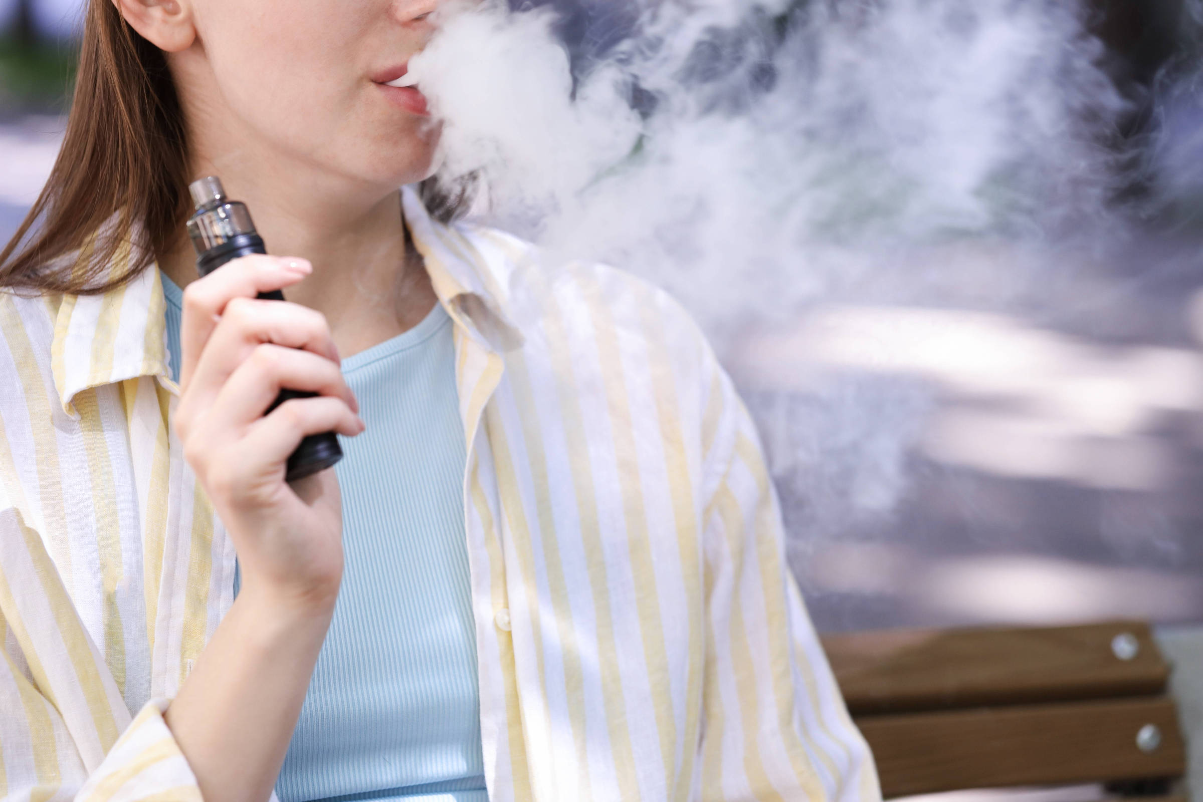 Pessoa segurando cigarro eletrônico próximo à boca, exalando grande nuvem de vapor branco. Veste camiseta azul clara e camisa listrada amarela e branca, sentada em banco de madeira com fundo desfocado.