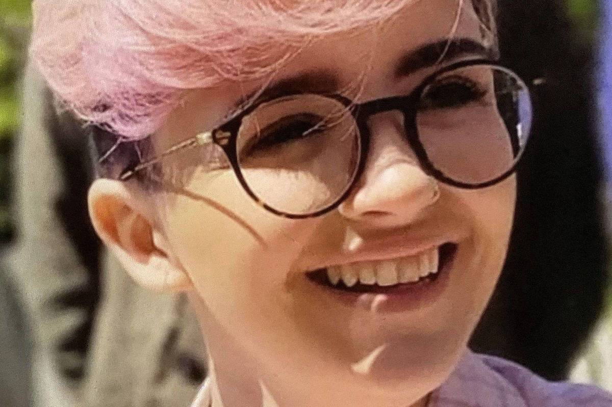 Pessoa jovem com cabelo tingido de rosa claro, usando óculos de armação redonda preta, sorrindo. Fundo desfocado com vegetação e sombra.