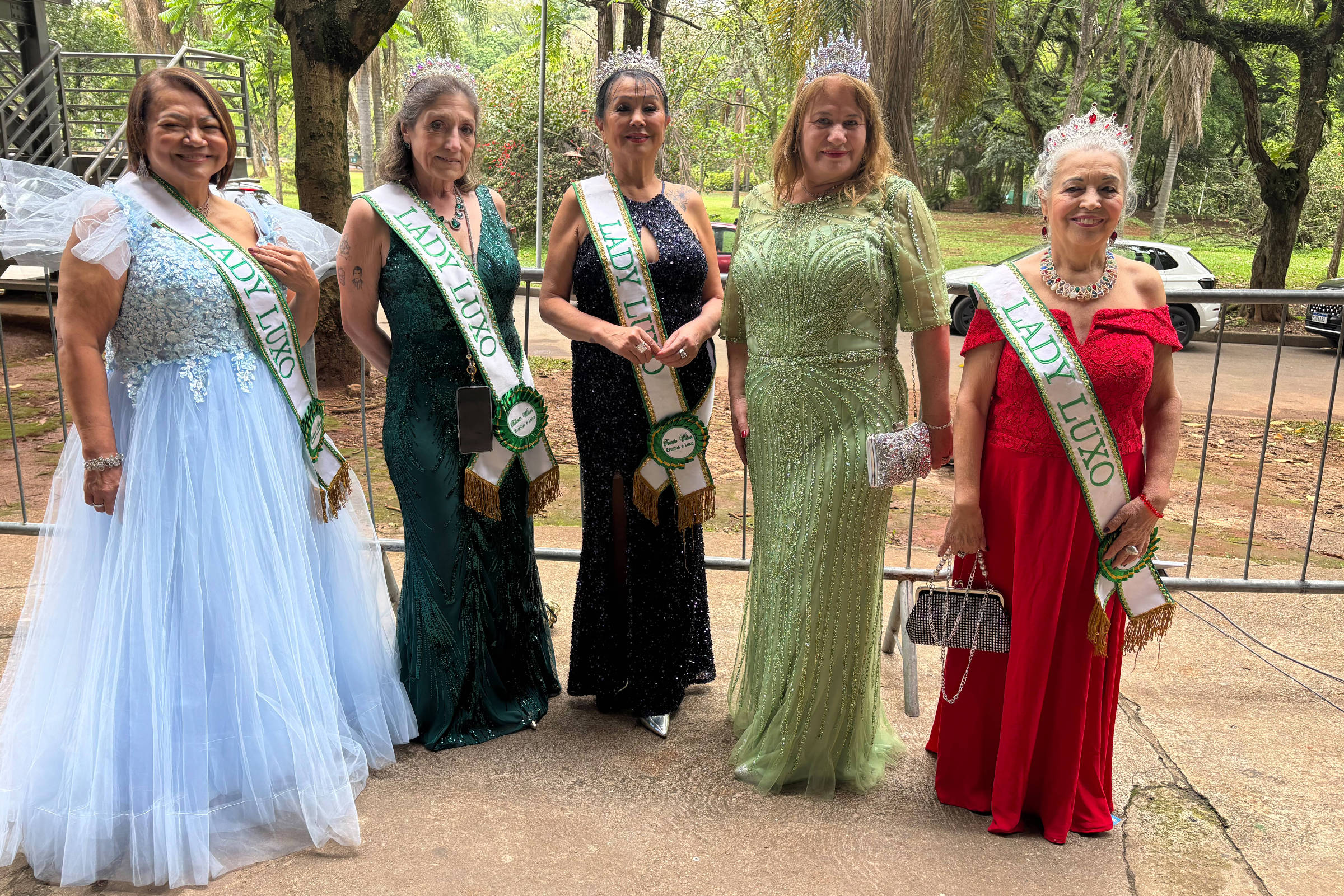 Cinco mulheres posam lado a lado em área externa com árvores ao fundo. Elas vestem vestidos longos de gala em cores azul claro, verde escuro, preto, verde claro e vermelho. Quatro delas usam faixas verdes com a inscrição