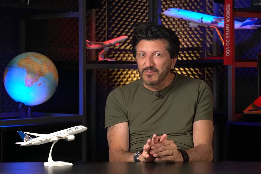 Homem de camiseta verde sentado à mesa em estúdio com iluminação escura. Ao fundo, prateleiras exibem dois modelos de aviões, um globo terrestre iluminado e um disco de vinil vermelho com capa da banda Queen.