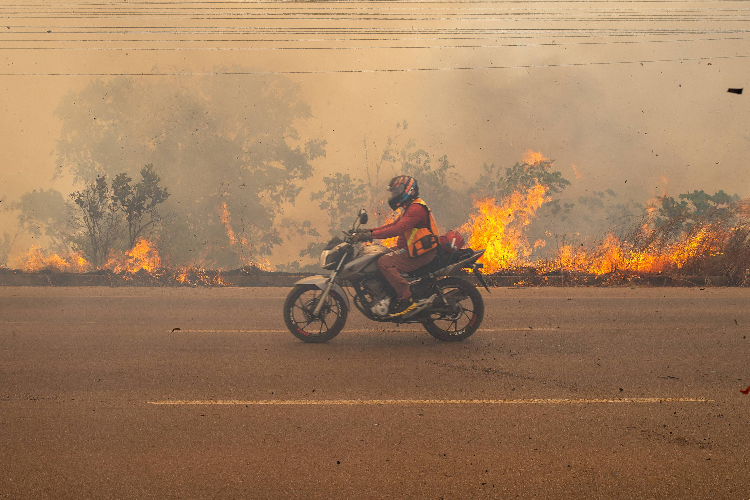 Motociclista com capacete e colete vermelho trafega em estrada asfaltada enquanto fogo consome vegetação seca ao fundo. Fumaça densa cobre o céu e árvores parcialmente queimadas são visíveis.