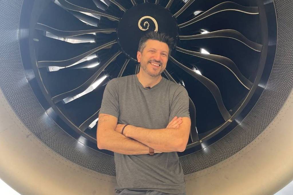 Homem de camiseta cinza e braços cruzados está em pé diante de uma grande turbina de avião com hélices visíveis ao fundo.