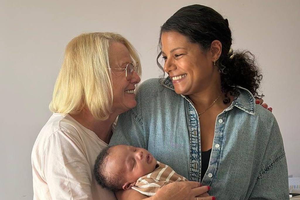 Duas mulheres estão próximas, sorrindo uma para a outra. Uma delas segura um bebê recém-nascido enrolado em manta listrada. O fundo é neutro, com parede clara e planta ao lado.