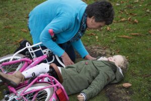 Mulher de cabelo curto e suéter azul ajoelhada ao lado de criança de casaco verde caída na grama, bicicleta rosa caída ao lado.
