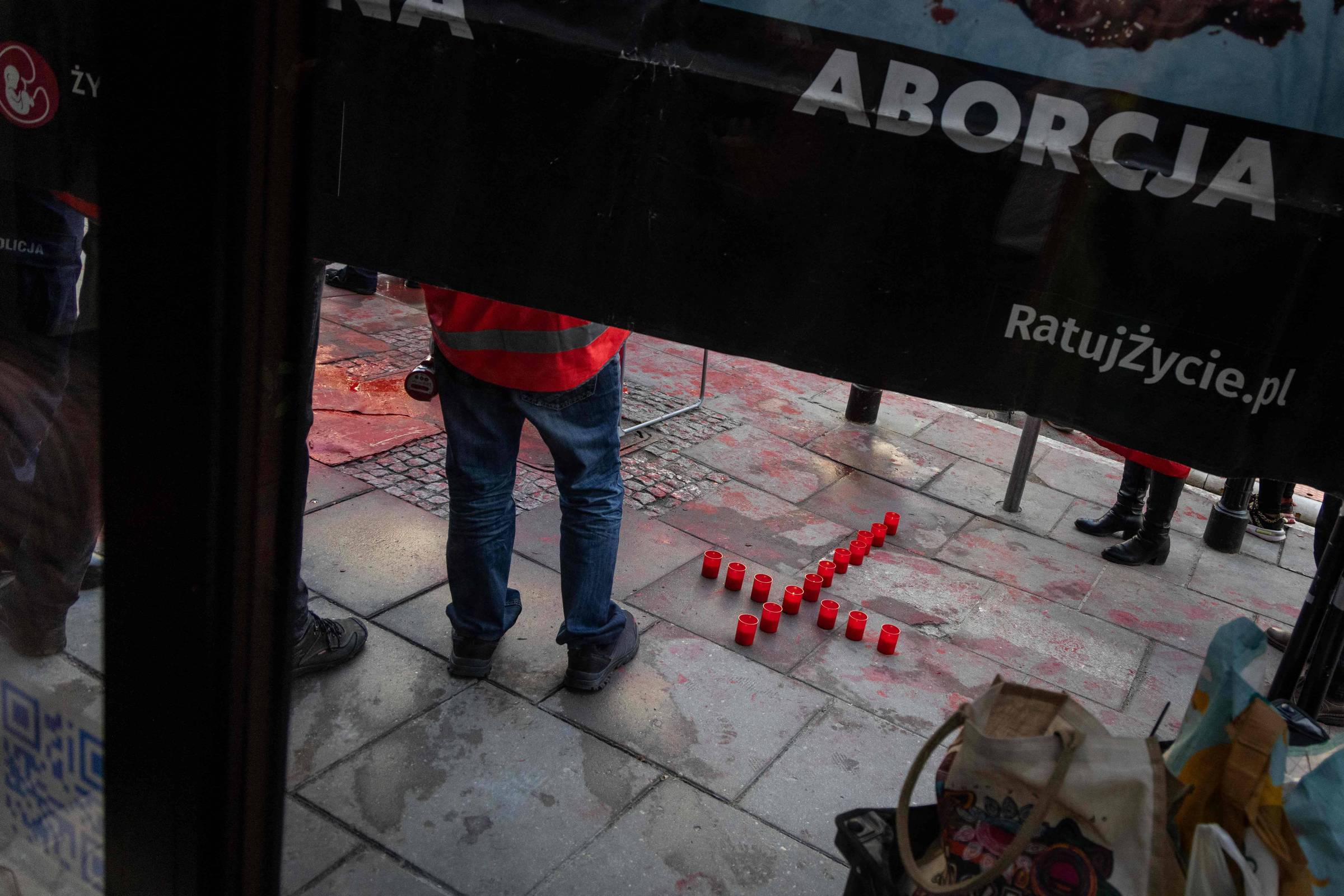 Velas vermelhas dispostas em forma de cruz no chão de pedra. Pessoas em pé ao redor, visíveis da cintura para baixo, com roupas casuais. Parte de faixa preta com texto em polonês aparece na parte superior da imagem.