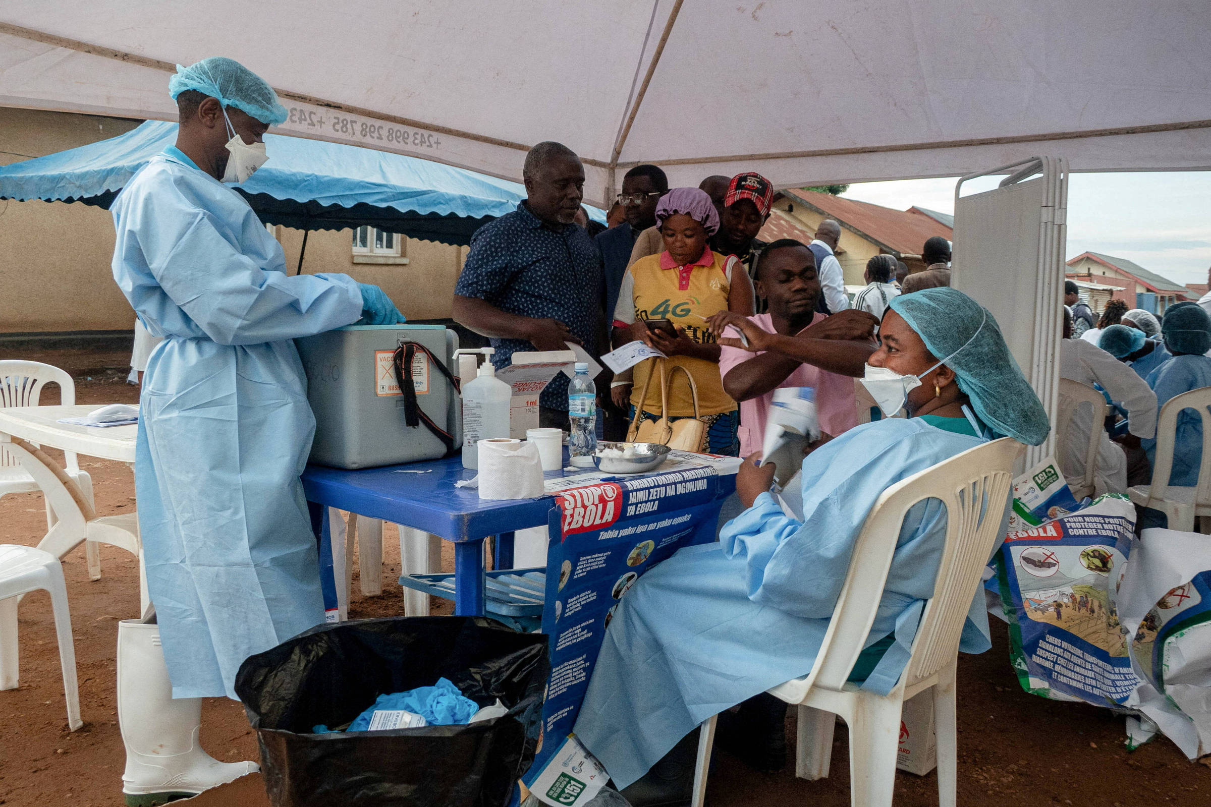 Profissionais de saúde com equipamentos de proteção aplicam vacinas em pessoas sob tenda. Indivíduos aguardam atendimento em fila próxima a mesa com insumos médicos.